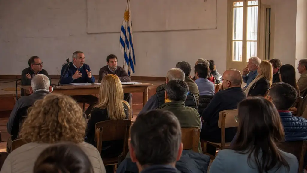 Asamblea territorial con bandera uruguaya: panel y vecinos debaten educación. Ilustración editorial · Nación UY
