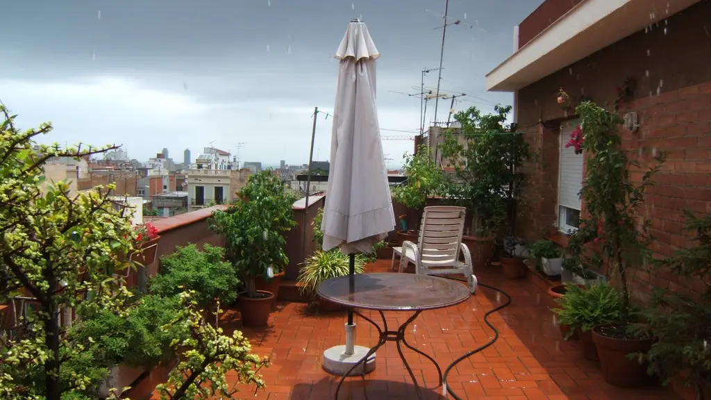 Lluvia sobre una azotea con plantas y una sombrilla cerrada, con edificios de Barcelona bajo un cielo gris al fondo. Foto: Literat Tours / Wikimedia Commons (CC BY-SA 3.0) (archivo)