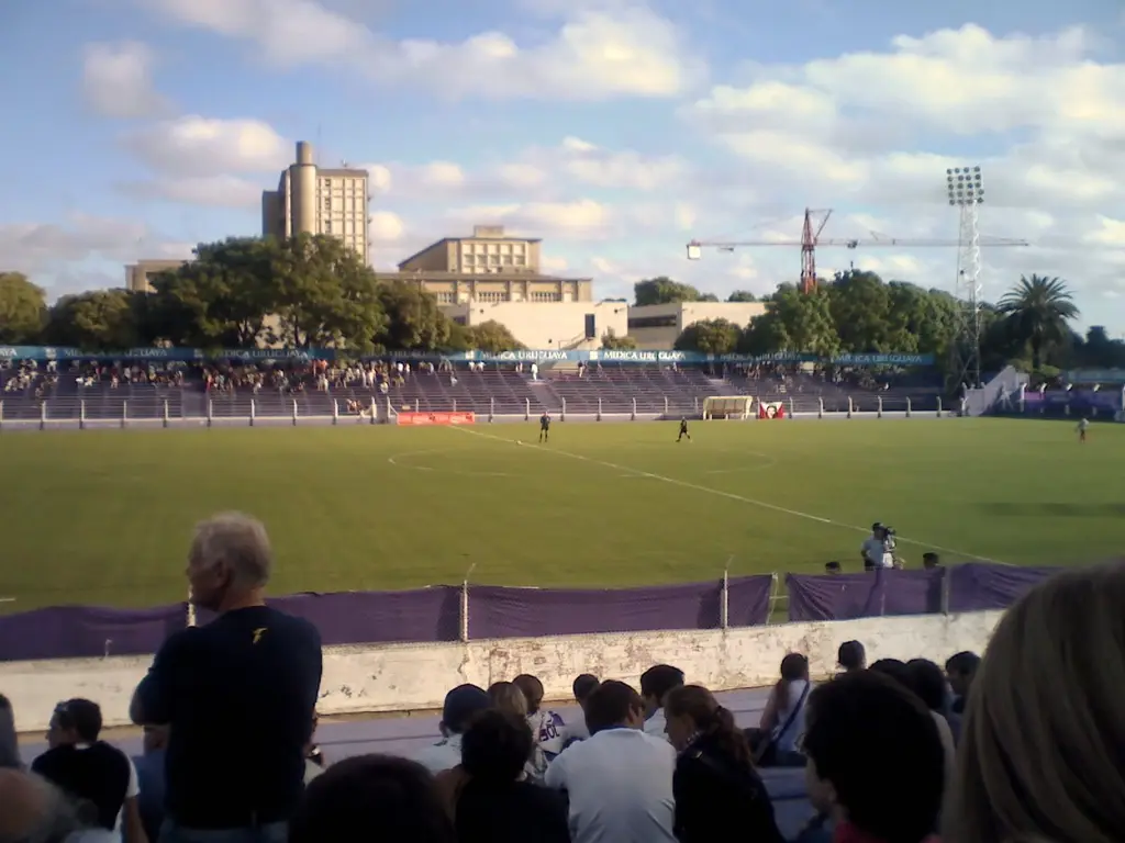 Hinchas en la tribuna del estadio Luis Franzini antes de un partido del fútbol uruguayo, con la cancha y edificios de fondo. Foto: Sebaclemente9 / Wikimedia Commons (CC BY-SA 4.0) (archivo)